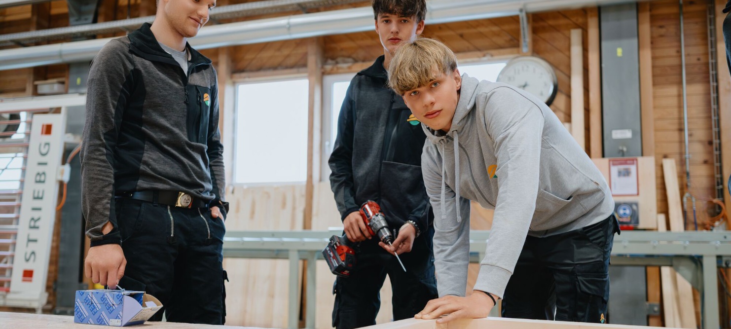 Eine Gruppe mit 3 Lernenden. Eine Person schaut in die Kamera, 2 Personen schauen auf das Werkstück.
